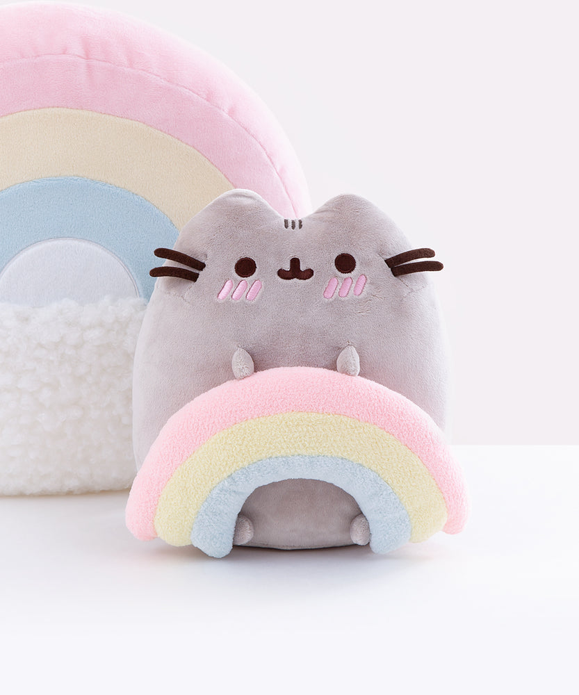 Pusheen sitting upright and blushing, two nub paws holding a plush rainbow and two nub paws at the bottom facing forward. The back of the Rainbow Pushen Plush Pillow is visible in the background. Pusheen’s Blush marks are three pink embroidered angled stripes directly underneath Pusheen’s eyes, and the rainbow is a pastel pink, yellow and blue.