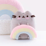 Pusheen sitting upright and blushing, two nub paws holding a plush rainbow and two nub paws at the bottom facing forward. The back of the Rainbow Pushen Plush Pillow is visible in the background. Pusheen’s Blush marks are three pink embroidered angled stripes directly underneath Pusheen’s eyes, and the rainbow is a pastel pink, yellow and blue.