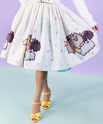 Full view of front of the Pusheenicorn Magical Jewel Skirt worn by a model who is wearing yellow shoes and standing in front of a light purple background. The skirt is held out to one side so the skirt falls away from the body to show off the repeating print pattern. 