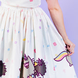 Closeup view if printed Pusheenicorn pattern skirt pattern. White skirt is dotted with pastel pink, purple, blue and yellow magical jewels with a repeating border pattern of Pusheenicorn carrying a pink cupcake in her mouth while walking alongside her and friends. 