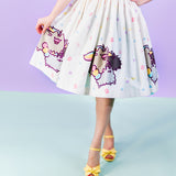 Front view of white retro-inspired skirt featuring a multicolor pastel confetti pattern and a repeating graphic of Pusheenicorn, Stormicorn and Pipicorn at the bottom hem. The model’s hand holds skirt fabric away from the body while the other hand is placed at the hip. Model is wearing yellow shoes with bows and standing in front of a purple background and light blue floor.  