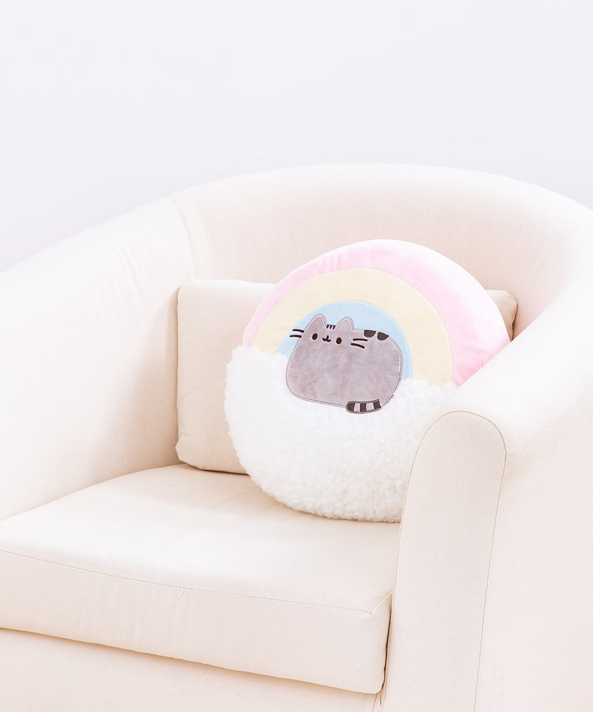 The Rainbow Pusheen Plush Pillow resting in a cream arm chair in front of a white background. The pillow takes up roughly ¾ of the back of the chair.