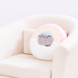 The Rainbow Pusheen Plush Pillow resting in a cream arm chair in front of a white background. The pillow takes up roughly ¾ of the back of the chair.