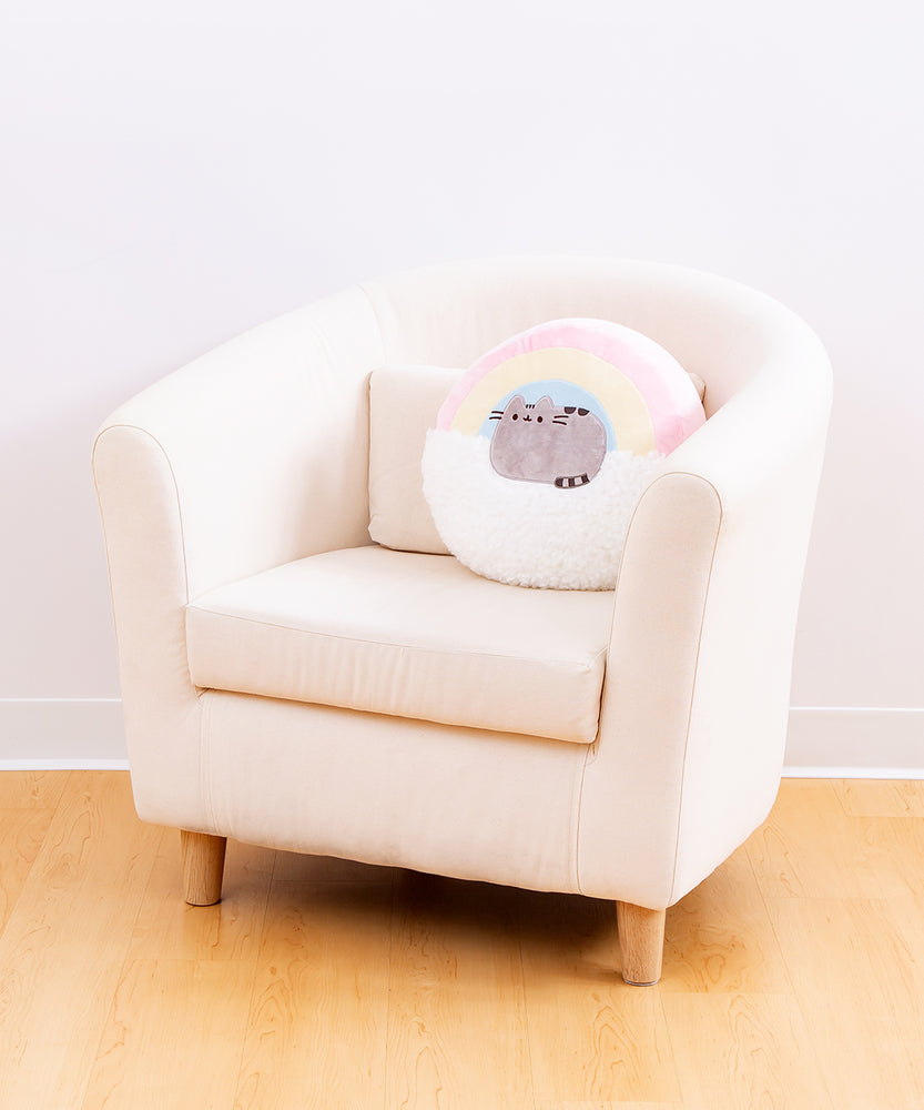 Full view of the Rainbow Pusheen Plush Pillow resting in a cream arm chair, on top of a wooden floor in front of a white background.