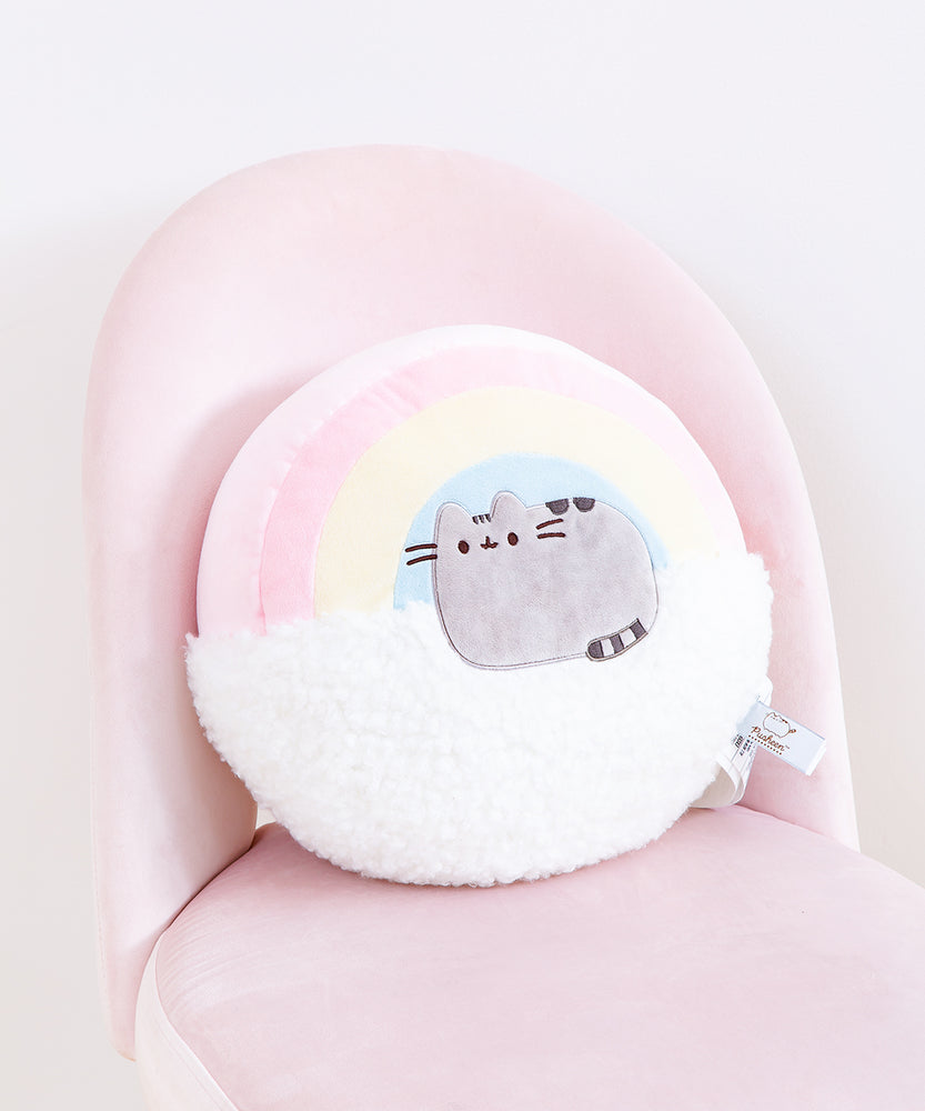 A circular pillow, half a pastel red, yellow and blue rainbow, half a fuzzy white cloud, with Pusheen sitting in loaf position right in the middle. The item tags for this item are visible in the bottom right corner of the pillow. The pillow is on top of a pink chair in front of a white background. 