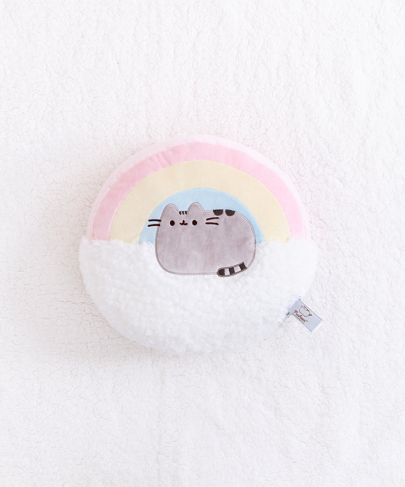 Overhead view of the Rainbow Pusheen Plush Pillow on top of a fuzzy white blanket. The item tag in the bottom right edge is visible.