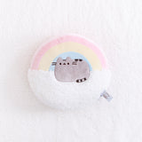 Overhead view of the Rainbow Pusheen Plush Pillow on top of a fuzzy white blanket. The item tag in the bottom right edge is visible.