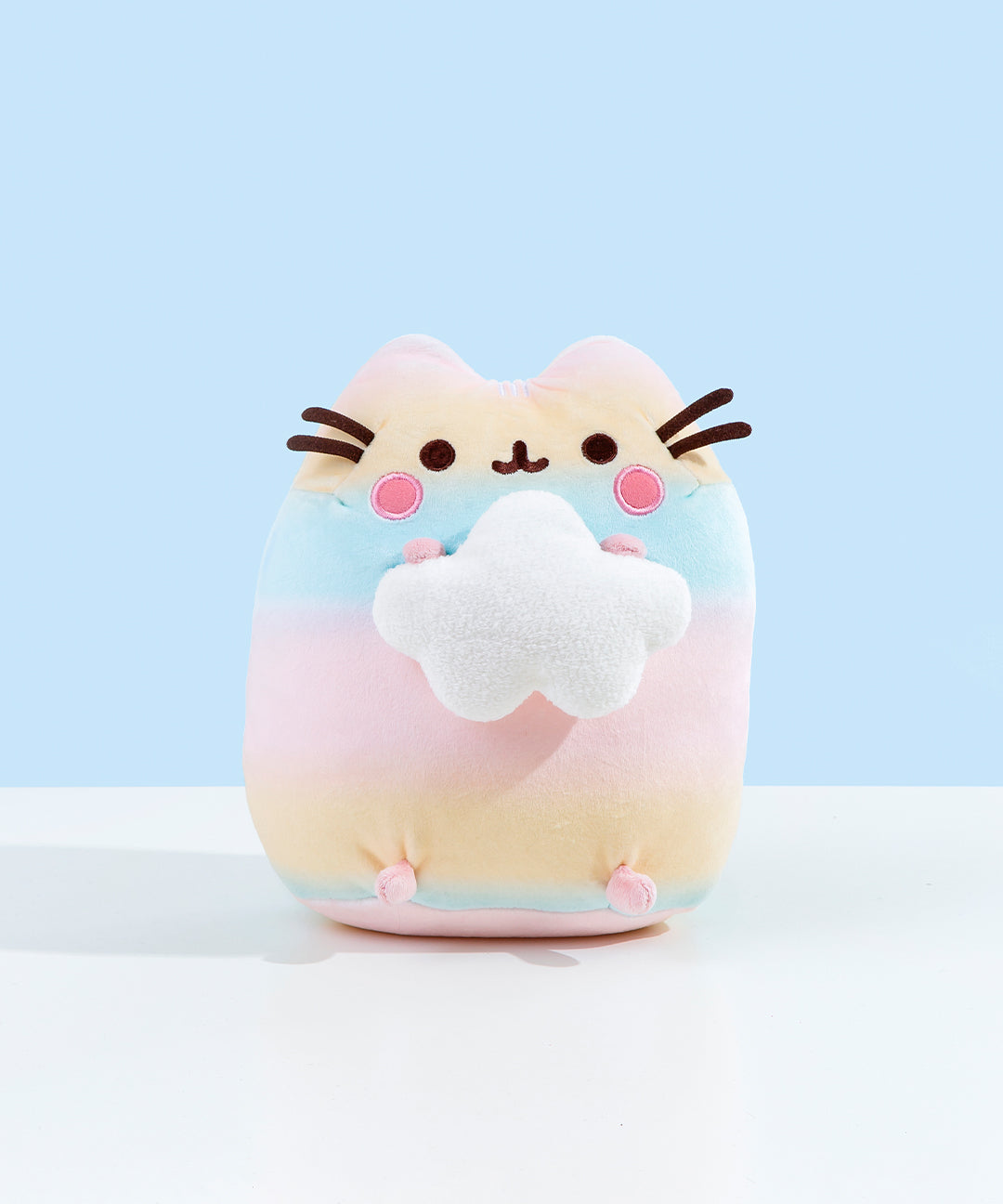 Pusheen colored in a pastel gradient, alternating between pink, yellow and blue. Pusheen is sitting upright, holding onto a plush white cloud in the shape of a star. Pusheen's stripes, eyes, and a round pink plush by her whiskers are embroidered into the plush. The whiskers are made of dark brown felt and point outwards.