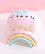 Rainbow Pusheen plush is angled on top of a pink, yellow, and blue pastel wooden rainbow. The plush’s paw details are yellow.  