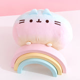 Rainbow Pusheen plush is angled on top of a pink, yellow, and blue pastel wooden rainbow. The plush’s paw details are yellow.  