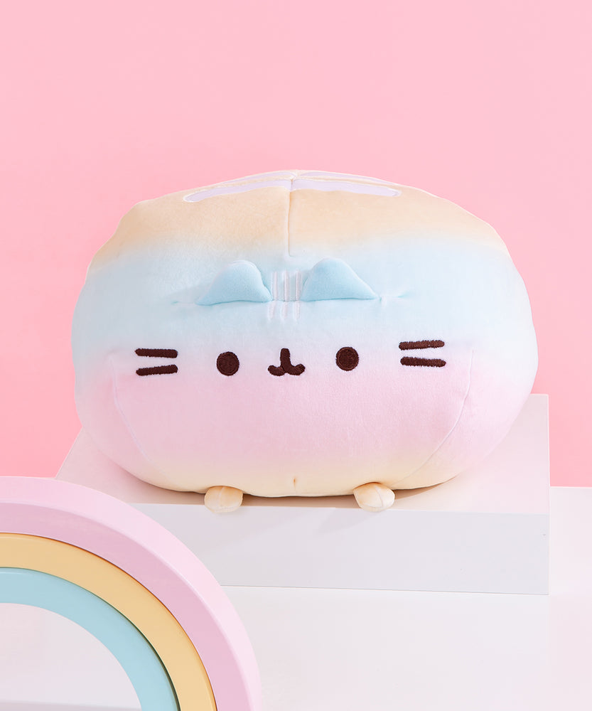 Front view of the Rainbow Round Pusheen Squisheen sitting on top of a white pedestal. A pink, yellow, and blue pastel rainbow sits in front of the plush. The yellow, blue, and pink pastel plush sits on four paws, two of which can be seen in this view.  
