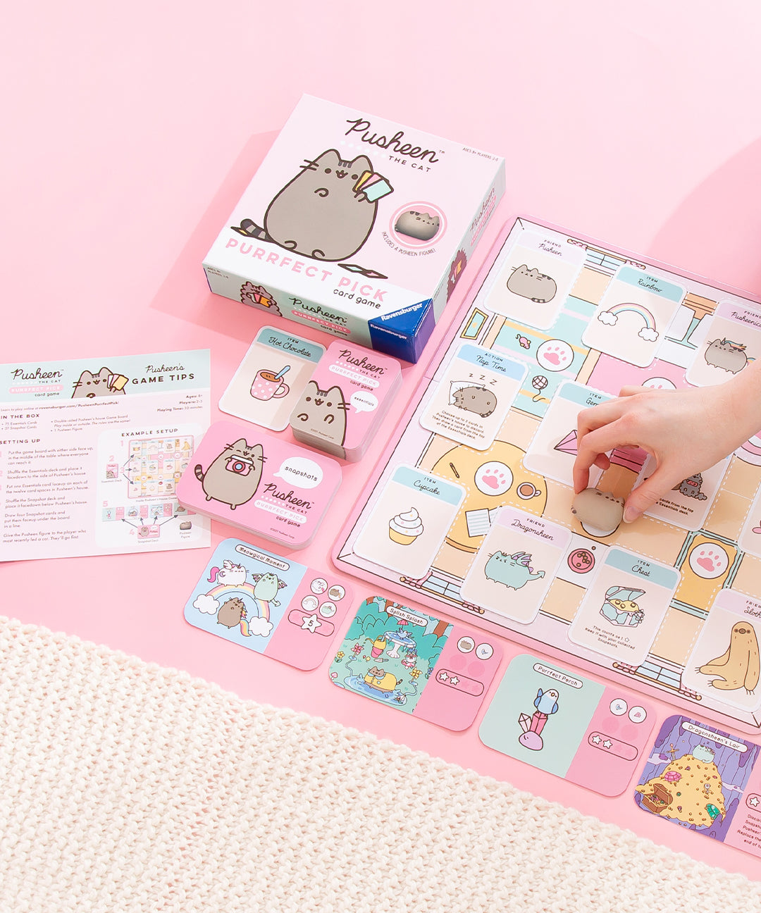 The contents of the Card Game neatly laid out on top of a pink floor, a model’s hand from the right picking up the Pusheen mini figurine. The contents of the game include a main board featuring an illustration of a living room, with various single cards resting on top. Around the board is the game’s box, a pile of cards stacked vertically and horizontally, four single cards with illustrations at the bottom, and the instruction sheet on the leftmost side.