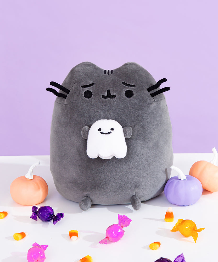 Front view of Pusheen Scaredy Cat Squisheen. The squisheen shows Pusheen in dark grey with a worried expression on her face. Her black whiskers and dark grey paws extend off her body. Pusheen holds a smiling white ghost in her front paws. The Squisheen is sitting on a white surface surrounded by purple, orange, and pink pumpkins and candies.  