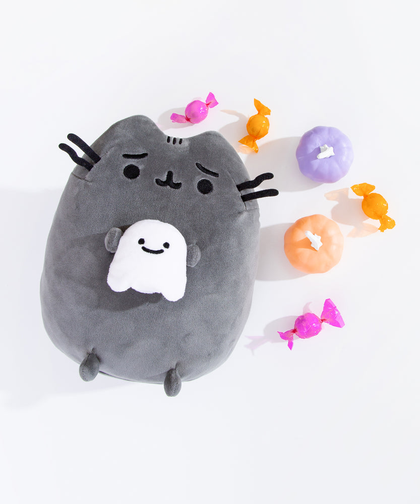 Pusheen Scaredy Cat Squisheen Plush lays on a white surface. The dark grey plush is holding a small smiling white ghost in her front paws. The plush is surrounded by pink and orange candies as well has purple and orange pumpkins.