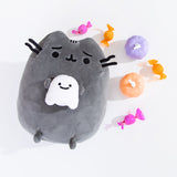 Pusheen Scaredy Cat Squisheen Plush lays on a white surface. The dark grey plush is holding a small smiling white ghost in her front paws. The plush is surrounded by pink and orange candies as well has purple and orange pumpkins.