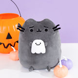 Halloween Squisheen Plush of Pusheen with a frightened expression holding a small white ghost in her paws. The Halloween plush is sitting on a white surface accompanied by orange and purple pumpkins and a jack-o-lantern Halloween candy bucket.
