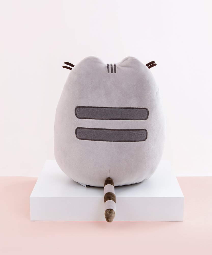 Back view of the Medium Sitting Squisheen, placed on top a square white pedestal in front of a light pink and white background. Two dark grey stripes are embroidered in Pusheen’s back, and a dark brown and light grey striped tail points out from the bottom center. Pusheen’s felt whiskers poke out from the front and are visible from the back. 
