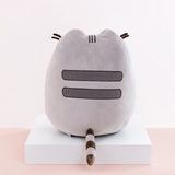 Back view of the Medium Sitting Squisheen, placed on top a square white pedestal in front of a light pink and white background. Two dark grey stripes are embroidered in Pusheen’s back, and a dark brown and light grey striped tail points out from the bottom center. Pusheen’s felt whiskers poke out from the front and are visible from the back. 