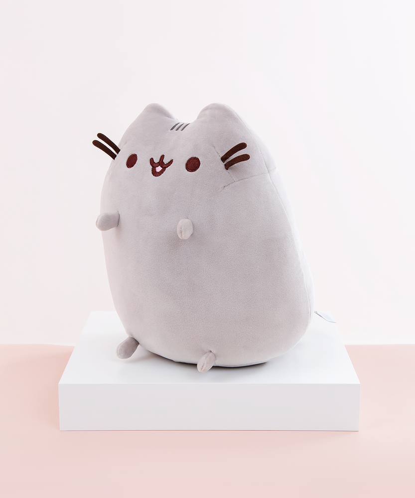 Quarter profile view of the Medium Sitting Squisheen, facing the left, placed on top a square white pedestal in front of a light pink and white background. The information tag is partially visible, located at the bottom right of Pusheen’s backside.