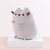 Quarter profile view of the Medium Sitting Squisheen, facing the left, placed on top a square white pedestal in front of a light pink and white background. The information tag is partially visible, located at the bottom right of Pusheen’s backside.