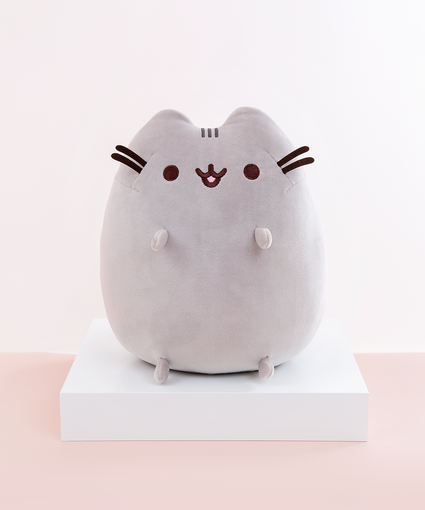 Pusheen sitting upright, with two nub paws up in the air and two nub paws at the bottom facing forward. Pusheen’s eyes and slightly open mouth are embroidered onto her face, and she has two felt whiskers poking out from each side of her face.  The medium sitting squisheen is on top of a white square podium, on top of a peach floor in front of a white background.