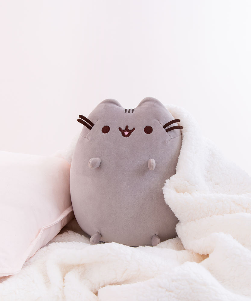 The Medium Sitting Squisheen Plush cozily seated besides a white pillow, with a white fuzzy blanket draped over her.