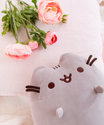Top view of the Medium Sitting Squisheen lying decadently on top of a fuzzy white blanket, head poking in from the bottom right to lie upon a pillow. Pink peonies are resting on top of the pillow as well in the upper left corner, right above Pusheen.