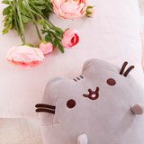Top view of the Medium Sitting Squisheen lying decadently on top of a fuzzy white blanket, head poking in from the bottom right to lie upon a pillow. Pink peonies are resting on top of the pillow as well in the upper left corner, right above Pusheen.