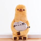 Front view of the Sloth plush holding the mini Pusheen plush in his arms, sitting on a white ledge in front of a white wall. The mini Pusheen plush is about the same width as Sloth’s body. 