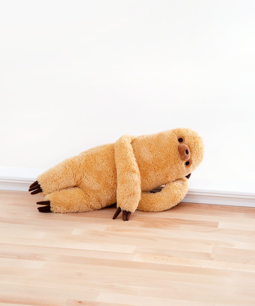 Sloth lounging on his side, right hand propping up his head and his left arm draped over his chest. Sloth is laid upon a wooden floor in front of a white background. He’s a real charmer, this lad.