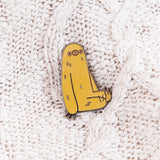 A pin of Sloth sitting on top of a white cable knit background. The pin features Sloth sitting upright, slightly slouched over, arms by his side. The line art is a reflective metal.