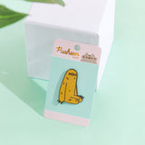 The Sloth pin in it’s packaging, leaning against a white pedestal on top a green floor, a plant leaf partially visible in the top left corner. The packaging is a round rectangular cardboard backing, a mint square in the middle and pink banners on the top and bottom. The top banner features the Pusheen Shop logo, and the bottom banner says ‘Sloth Pin’ in white.