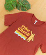 The Vibe Check tee laid out on a wooden floor at a slight angle, a pair of leaves above it.