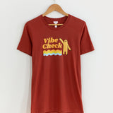 A maroon short sleeve tee hanging from a clothes hanger in front of a white background. The tee features a print of Sloth standing with his left arm raised, the words ‘Vibe Check’ to his left, and three squiggly lines in orange, olive and blue underneath the words.