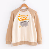 A full view of the top of the Pajama Set. The graphic features the words 'Smart Cookie', with a fuzzy embroidered image of Pusheen facing the left, pawing at a cookie on the floor in front of her. The sleeves, neck and bottom rib of the top are all tan, while the graphic is on top of the cream body.