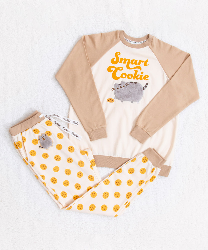 A full view of the tan and cream Pajama set top and bottom, partially folded on top of a fuzzy white background. The top has tan sleeves and a cream body with the graphic 'Smart Cookie' on it. The cream pants features a Pusheen patch and a repeating cookie pattern, with a tan waistband and cuffs.