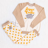 A full view of the tan and cream Pajama set top and bottom, partially folded on top of a fuzzy white background. The top has tan sleeves and a cream body with the graphic 'Smart Cookie' on it. The cream pants features a Pusheen patch and a repeating cookie pattern, with a tan waistband and cuffs.