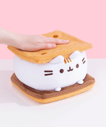 Gif of Pusheen S'mores Squisheen being smooshed by model hand. Pusheen the Cat takes the form of a s'mores treat. The top and bottom slabs of graham cracker-inspired plush is a soft textured light brown fabric.