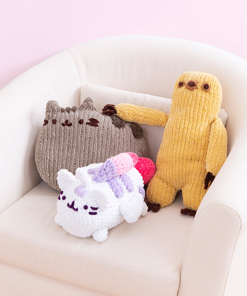 Three plush are arranged on a white chair. The smallest is the Super Pusheenicorn crocheted plush, behind which are Pusheen in a loaf pose and a Sloth knitted plush.