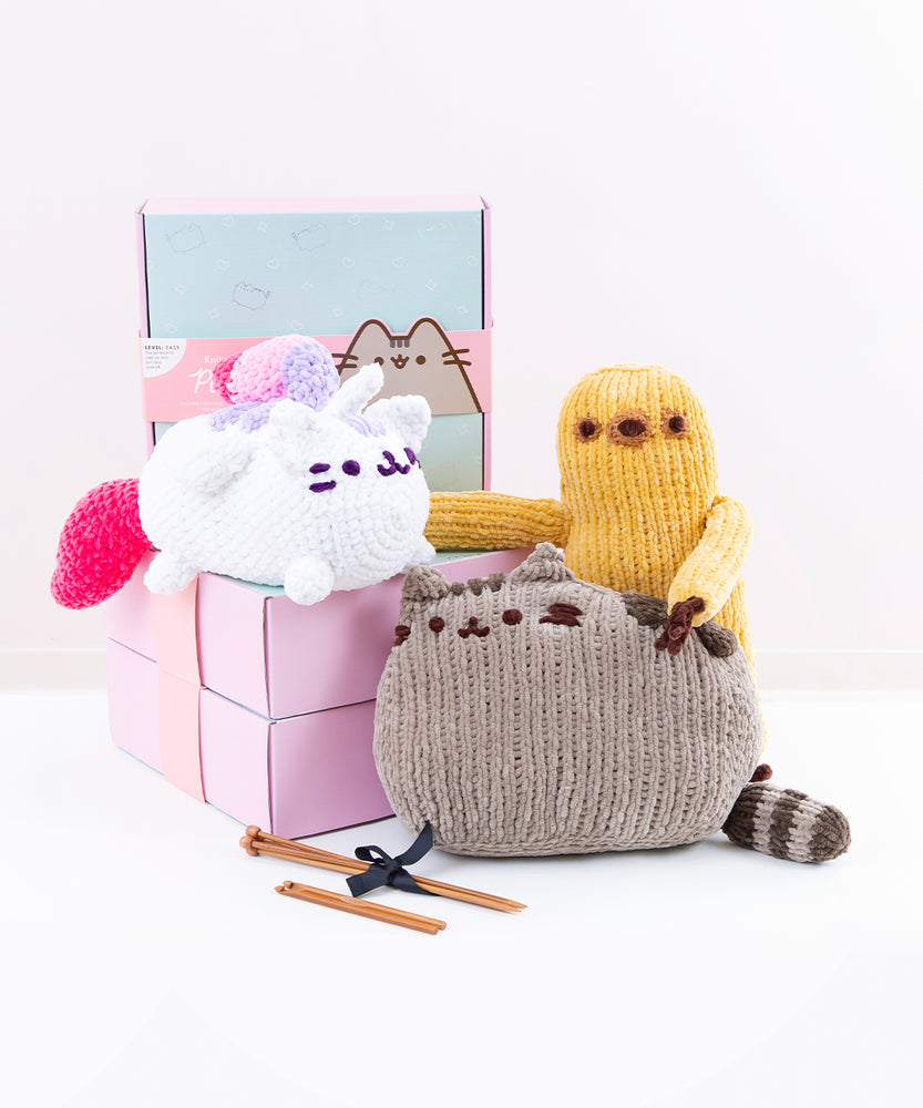 Three knitted or crocheted plush (Super Pusheenicorn, Pusheen, and Sloth) arranged on and in front of the square kit boxes with the knitting and crochet needles in front.