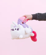 A hand from offscreen holds the Super Pusheenicorn crocheted plush by the mane in front of a pink background. The mane serves as a handle for the plush. 