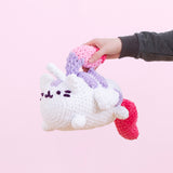 A hand from offscreen holds the Super Pusheenicorn crocheted plush by the mane in front of a pink background. The mane serves as a handle for the plush. 