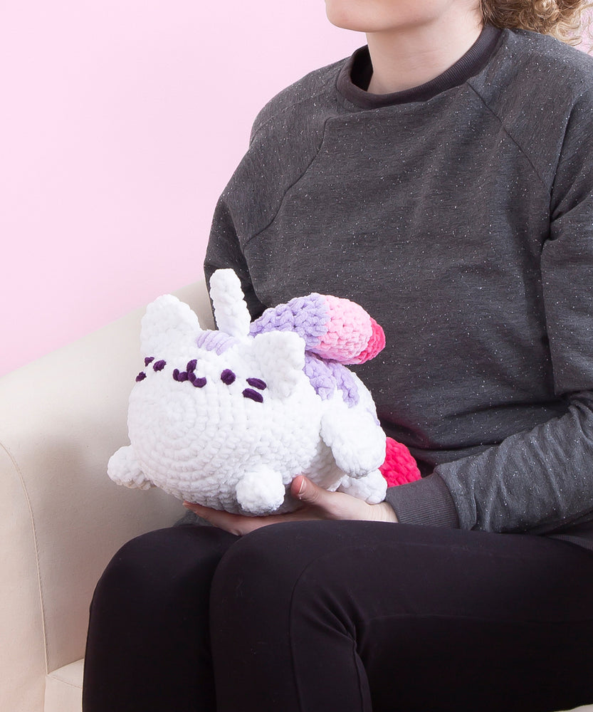 The crocheted Sloth, Pusheen, and Super Pusheenicorn plush being held in the arms of a figure with long curly hair.