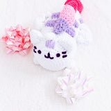 A completed Super Pusheenicorn Amigurumi, laying on her stomach on top of a fuzzy white blanket with shiny pink and white gift bows beside her. The completed crochet amigurumi features Super Pusheenicorn laying on her stomach in a log pose, her wings sticking out from the side and her purple, pink and dark pink mane going down her back. Some details such as her head stripes and face are sewn into the amigurumi.