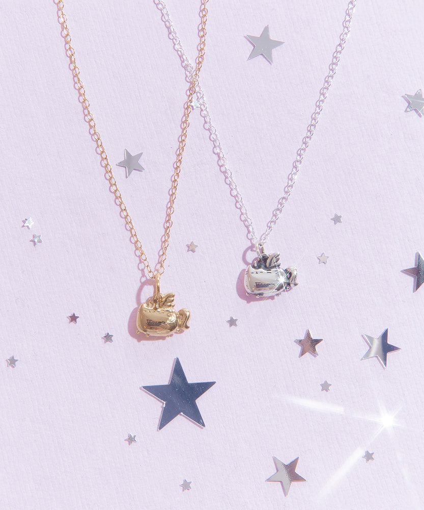 Two Super Pusheenicorn Charm Necklaces, one gold and one silver, lay next to each other on top of a light purple background, surrounded by reflective silver stars. The Super Pusheenicorn charm hangs from her wings and has her eyes closed dramatically, making it look as if she’s mid flight.  