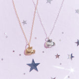 Two Super Pusheenicorn Charm Necklaces, one gold and one silver, lay next to each other on top of a light purple background, surrounded by reflective silver stars. The Super Pusheenicorn charm hangs from her wings and has her eyes closed dramatically, making it look as if she’s mid flight.  