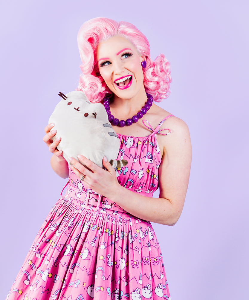 Front view of smiling model wearing Super Pusheenicorn Dreamer Dress while holding a medium-size plush of Pusheen the Cat in their hands.  