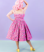 Back view of the dreamy pink dress shows off the pleating at the top of the skirt. Model is wearing yellow shoes and a pink wig while standing with one hand on the hip and one hand on the head. 