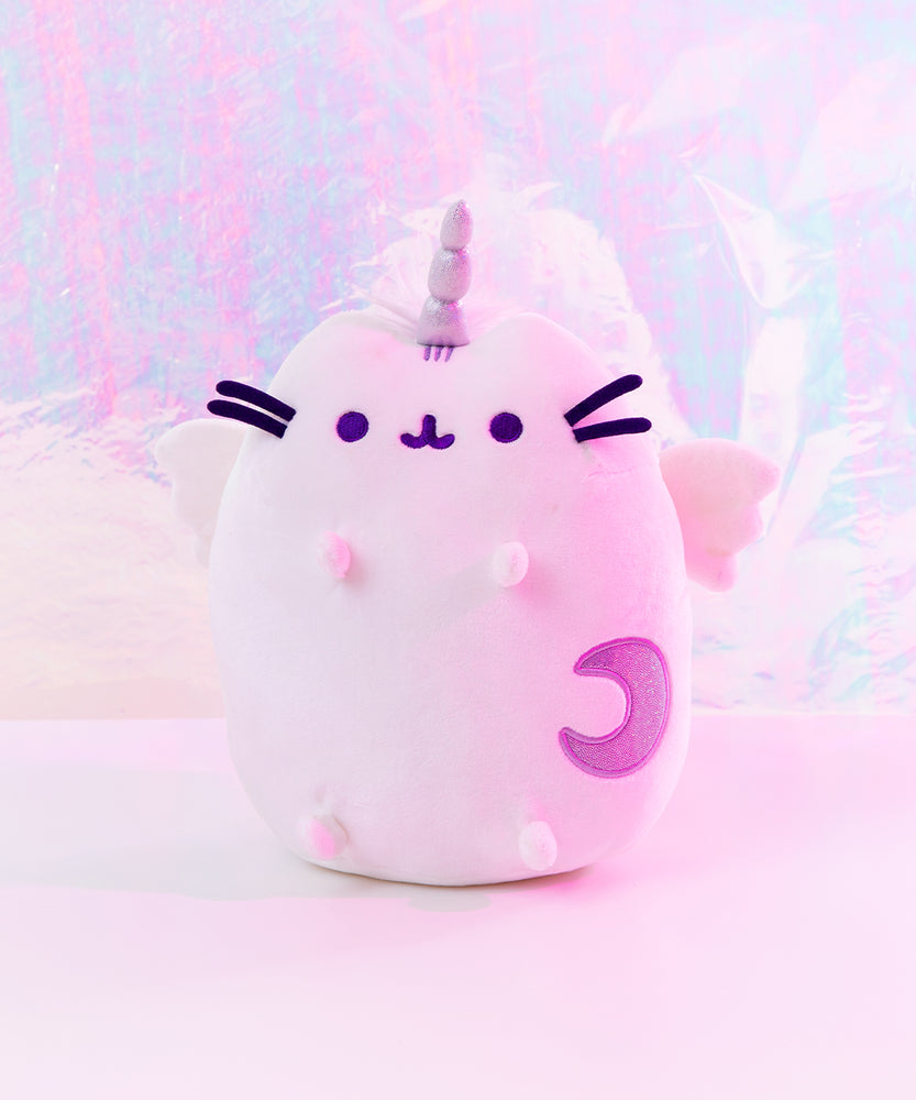 The Super Pusheencorn plush in front of a magical fluorescent background, bathed in a magenta light. The Super Pusheenicorn plush reflects the light around her and appears light pink as a result, her Moon mark appearing to be a darker purple.