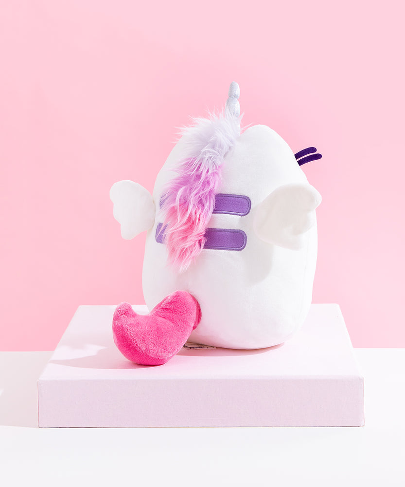 Back view of the Super Pusheenicorn Plush on top of a white square pedestal, in front of a pink background. There is a multicolor mane directly behind Super Pusheenicorn’s horn, colored light purple, purple and pink, going over Pusheen’s Back stripes and ending at the bottom stripe. Pusheen’s back stripes are embroidered in a dark purple, and besides them are two plush wings. Super Pusheenicorn has a solid pink plush tail that curves upwards.
